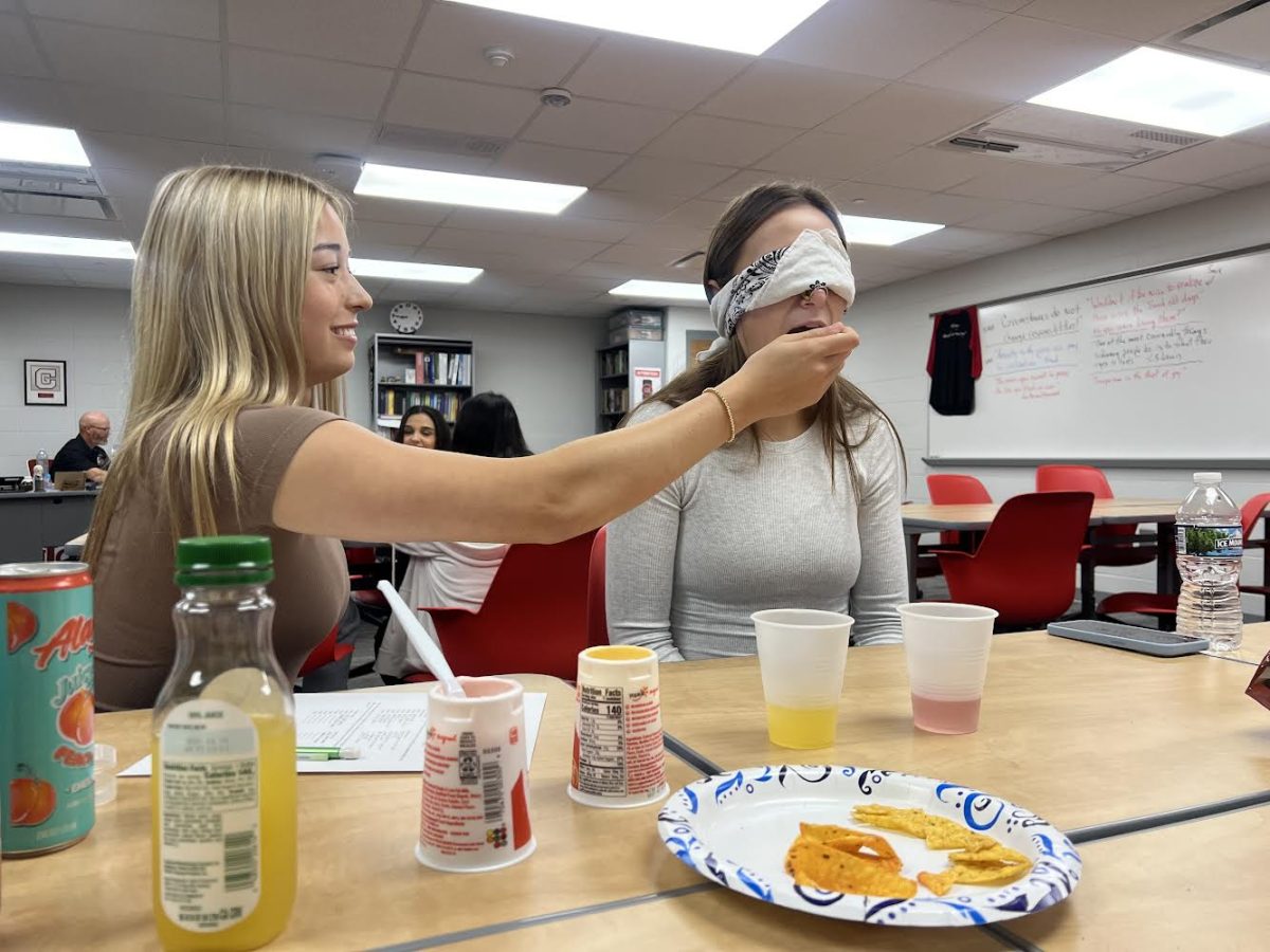 Senior Hannah Vassar feeds a blindfolded senior Addie Crowe different foods and drinks for AP psychology. Each AP psychology class had one student from each table taste foods and drinks without sight or smell to see just how good their senses and taste buds are. “I knew when you fed me chips compared to yogurt,” Crowe said. “Those were the easiest to identify. This activity helped me establish that I don’t have very strong taste buds to tell the difference between foods.”