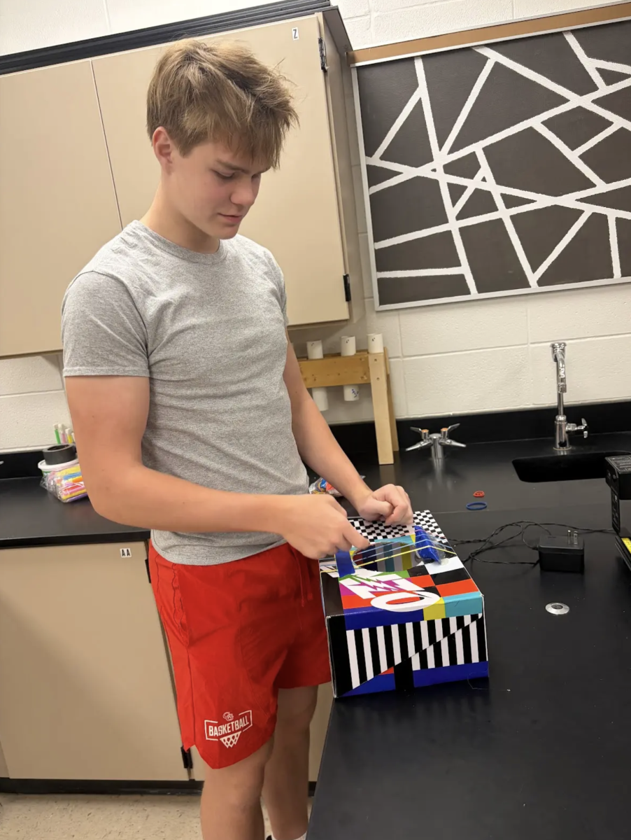 Junior Rhys Oliver plucks the strings of his shoebox guitar during Mr. Werner’s Physics I class. Students designed and made musical instruments out of simple everyday items and tested them to see if they produced the right pitch. “We cut out a hole in the middle of our shoebox, and then we put rubber bands around it to act as strings just like a guitar,” Oliver said. “We used different thicknesses of rubber bands and propped them up so they were tight which allowed them to play the pitches that we wanted.”